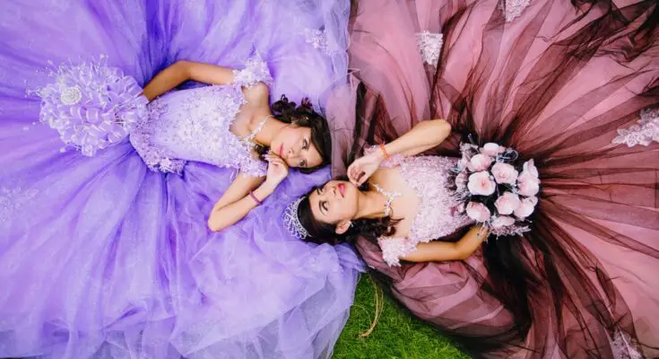 Two teenage girls in quinceañera dresses, one lilac and one dusty rose, lay in the grass and look at once another.