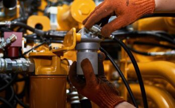 A pair of hands wearing dirty orange work gloves screwing a metal part onto a yellow hydraulic pipe system.