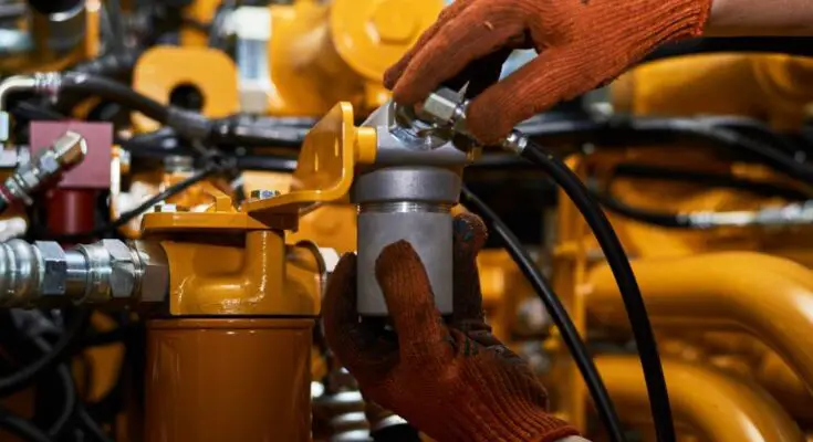 A pair of hands wearing dirty orange work gloves screwing a metal part onto a yellow hydraulic pipe system.