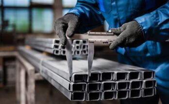 A worker wearing a blue jumpsuit and dark gray gloves uses a tool to measure the width of a rectangular metal tube.