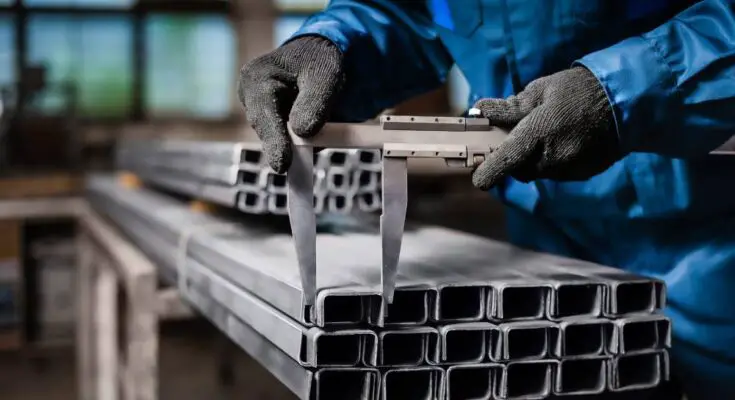 A worker wearing a blue jumpsuit and dark gray gloves uses a tool to measure the width of a rectangular metal tube.