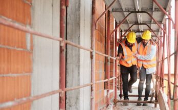 A construction worker in a white, long-sleeve shirt helping his coworker in a black t-shirt walk through a construction site.