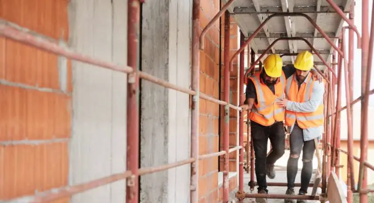 A construction worker in a white, long-sleeve shirt helping his coworker in a black t-shirt walk through a construction site.