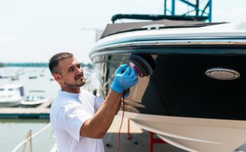 A man using an orbital polisher to shine a boat, set against a scenic marina backdrop with other boats and clear blue skies.