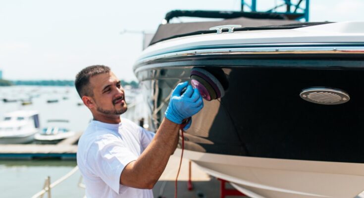 A man using an orbital polisher to shine a boat, set against a scenic marina backdrop with other boats and clear blue skies.