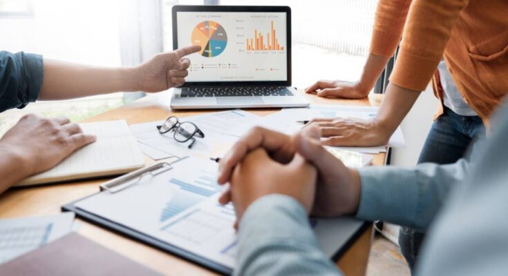 A diverse group of coworkers gathered around a table and focused on a laptop screen displaying charts and graphs.