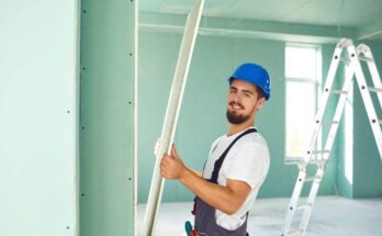 A construction worker installing plasterboard drywall inside a nearly complete building. A ladder stands in the background.