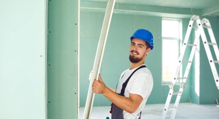 A construction worker installing plasterboard drywall inside a nearly complete building. A ladder stands in the background.