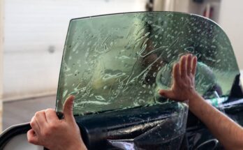 A person is applying a sheet of tint to a vehicle's driver's side window. A hand is smoothing the bubbles.