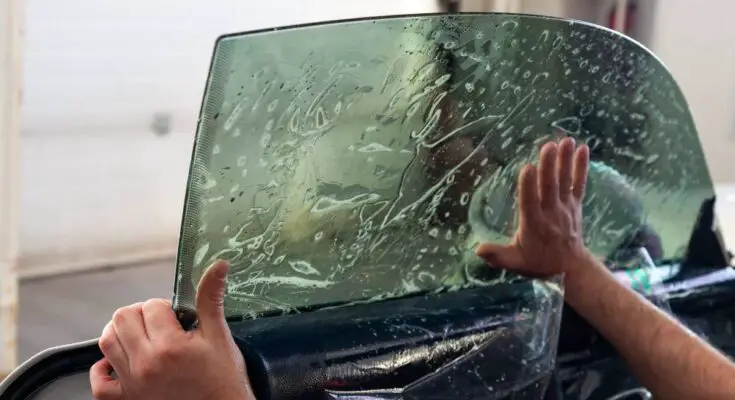 A person is applying a sheet of tint to a vehicle's driver's side window. A hand is smoothing the bubbles.