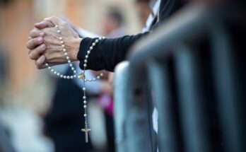 A woman’s hands laced together in prayer as she holds a white and gold Rosary. Past her is a blurred congregation.