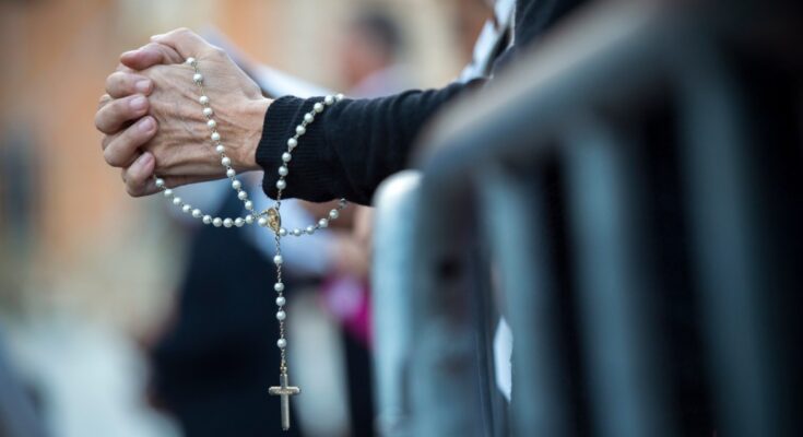 A woman’s hands laced together in prayer as she holds a white and gold Rosary. Past her is a blurred congregation.