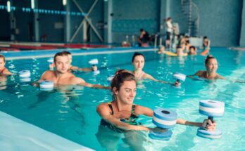 A group of six adults performing water aerobics in an indoor pool, extending their arms to the left with foam weights.