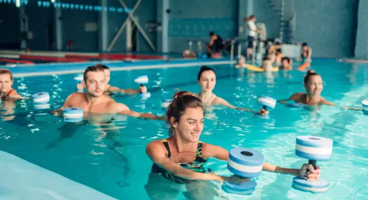 A group of six adults performing water aerobics in an indoor pool, extending their arms to the left with foam weights.