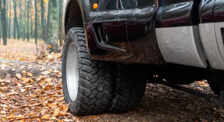 The large front driver's side wheel on an extra-large pickup truck is visible. The setting is an autumnal scene.