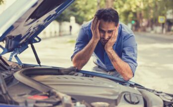 A man in a blue shirt holding his head in his hands and making a worried expression while looking at his car's engine.