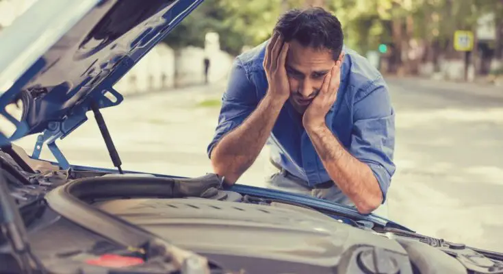 A man in a blue shirt holding his head in his hands and making a worried expression while looking at his car's engine.