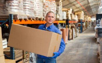 A man in a blue button-up shirt and jeans carrying a large cardboard box through an aisle in a warehouse.