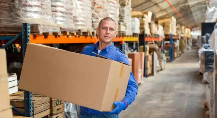 A man in a blue button-up shirt and jeans carrying a large cardboard box through an aisle in a warehouse.