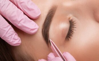 A woman reclines in a salon chair while a technician wearing pink gloves uses tweezers to shape and pluck her eyebrows.