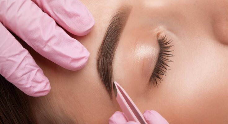 A woman reclines in a salon chair while a technician wearing pink gloves uses tweezers to shape and pluck her eyebrows.