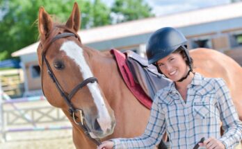 A avid horse rider wearing a riding helmet and holding onto the horse's nozzle. She is smiling and the horse is posed.