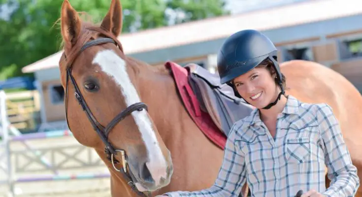 A avid horse rider wearing a riding helmet and holding onto the horse's nozzle. She is smiling and the horse is posed.