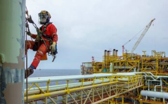 An oil rig worker on a hoist above the rig. The rig is offshore and the worker is wearing a helmet and protective gear.