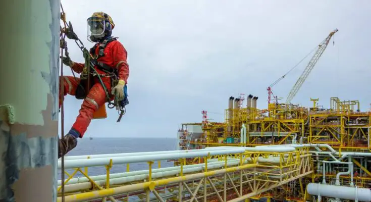 An oil rig worker on a hoist above the rig. The rig is offshore and the worker is wearing a helmet and protective gear.