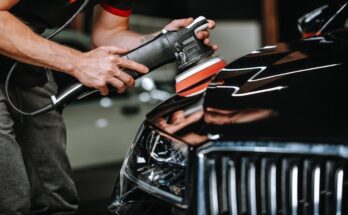 A man holding an orbital polisher against a sleek black car in a car studio. He is detailing and cleaning the car.