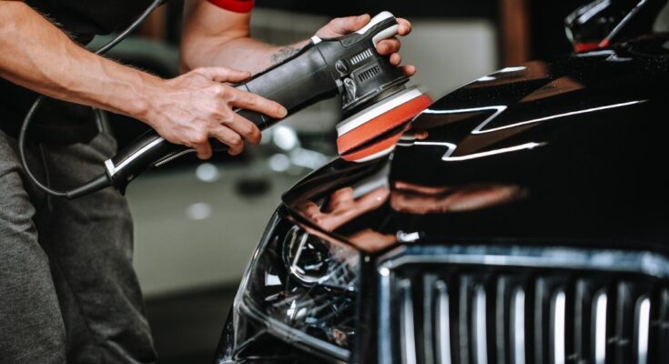 A man holding an orbital polisher against a sleek black car in a car studio. He is detailing and cleaning the car.