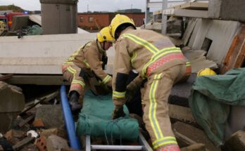 Two firefighters climb through rubble from a collapsed building with a ladder and a green blanket.