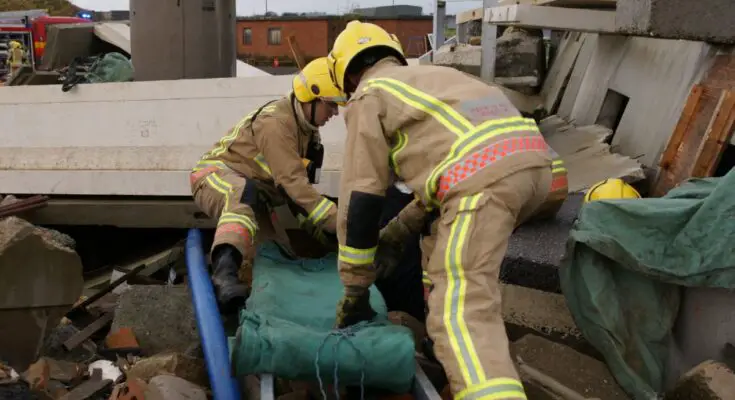 Two firefighters climb through rubble from a collapsed building with a ladder and a green blanket.