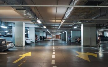 A large underground parking lot with a mostly monochromatic color scheme. Several parking spaces are occupied.