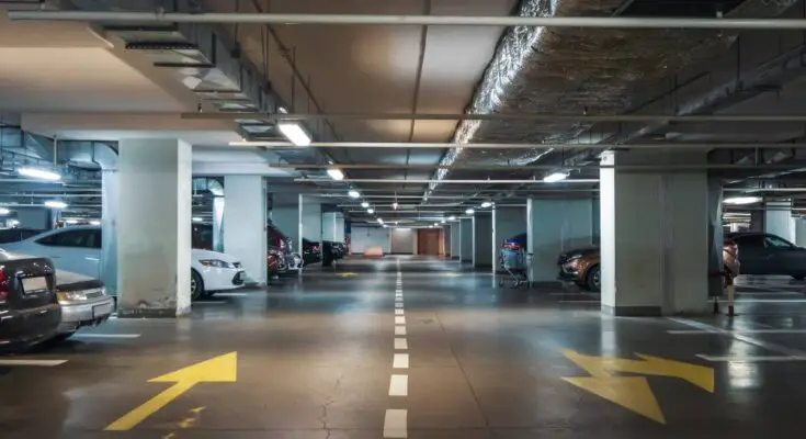 A large underground parking lot with a mostly monochromatic color scheme. Several parking spaces are occupied.