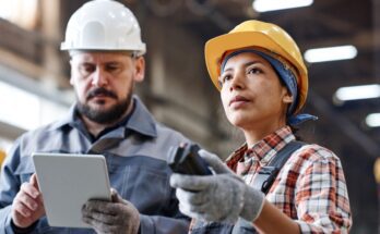 Two warehouse employees standing side-by-side. One is using a tablet to review stock and another is gazing at the warehouse.