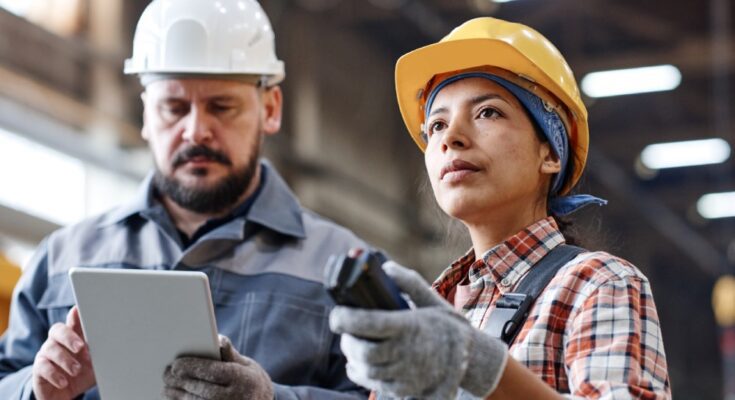 Two warehouse employees standing side-by-side. One is using a tablet to review stock and another is gazing at the warehouse.