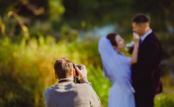 A photographer snaps pictures of a bridal couple outside in a green park. Only the photographer is in focus.