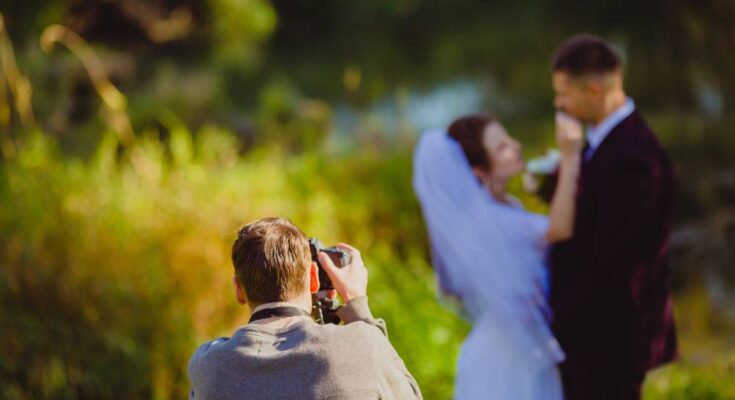 A photographer snaps pictures of a bridal couple outside in a green park. Only the photographer is in focus.