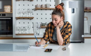 A woman in a black and yellow plaid shirt stands at a kitchen counter, writing in a notepad. Her hair is in a bun.