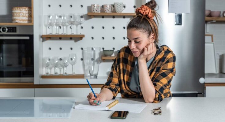 A woman in a black and yellow plaid shirt stands at a kitchen counter, writing in a notepad. Her hair is in a bun.