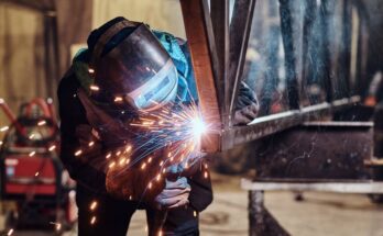 A metal fabricator wearing a welding mask and gloves as they hold a hand torch to cut a small metal beam.