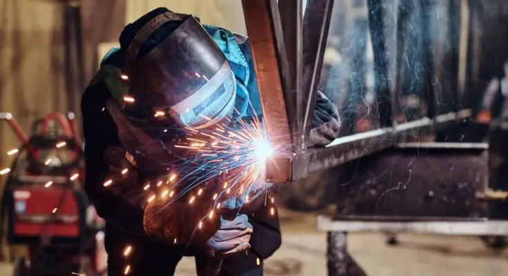 A metal fabricator wearing a welding mask and gloves as they hold a hand torch to cut a small metal beam.