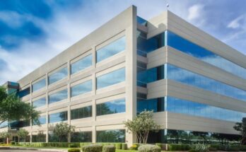 A gray, four-story commercial building stands high, surrounded by a blue sky. It has a parking lot, trees, and shrubs.