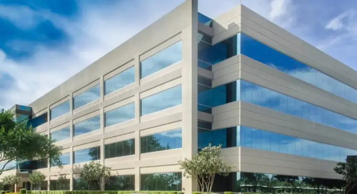 A gray, four-story commercial building stands high, surrounded by a blue sky. It has a parking lot, trees, and shrubs.