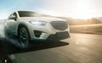 A close-up view shows a white car driving down the road with smoke coming off from the tires. The background is blurry.