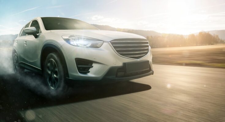 A close-up view shows a white car driving down the road with smoke coming off from the tires. The background is blurry.