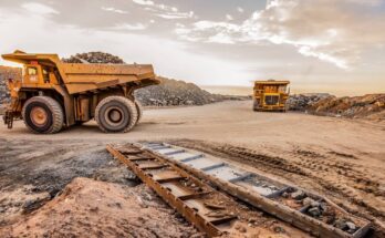 Two yellow mining dump trucks drive on a dirt road in a dusty landscape with their backs full of ore.