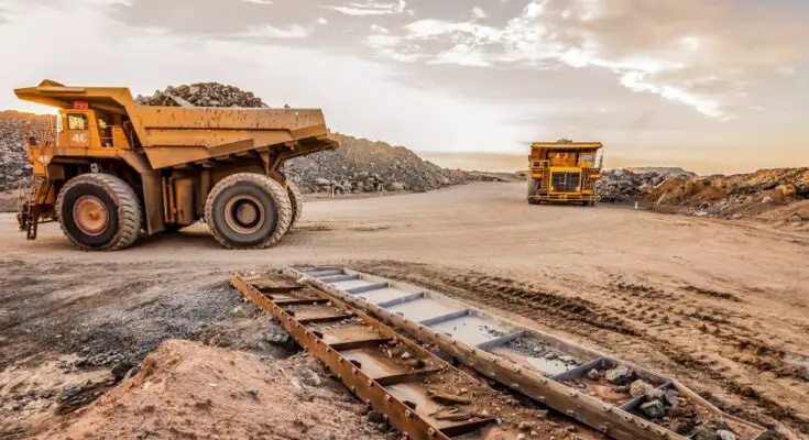 Two yellow mining dump trucks drive on a dirt road in a dusty landscape with their backs full of ore.