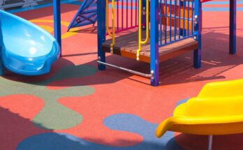 A playground with a blue slide and a yellow side, climbing equipment, and a rubber surface. The surface is red with blue designs.